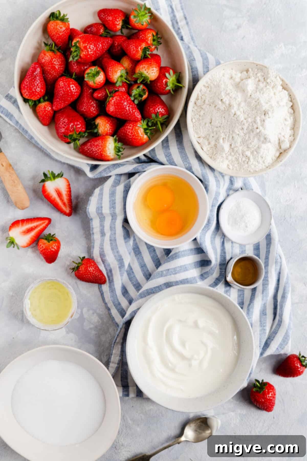 Top view of all the simple ingredients laid out for making the fresh strawberry yogurt cake: flour, sugar, eggs, yogurt, oil, vanilla, baking powder, and fresh strawberries.