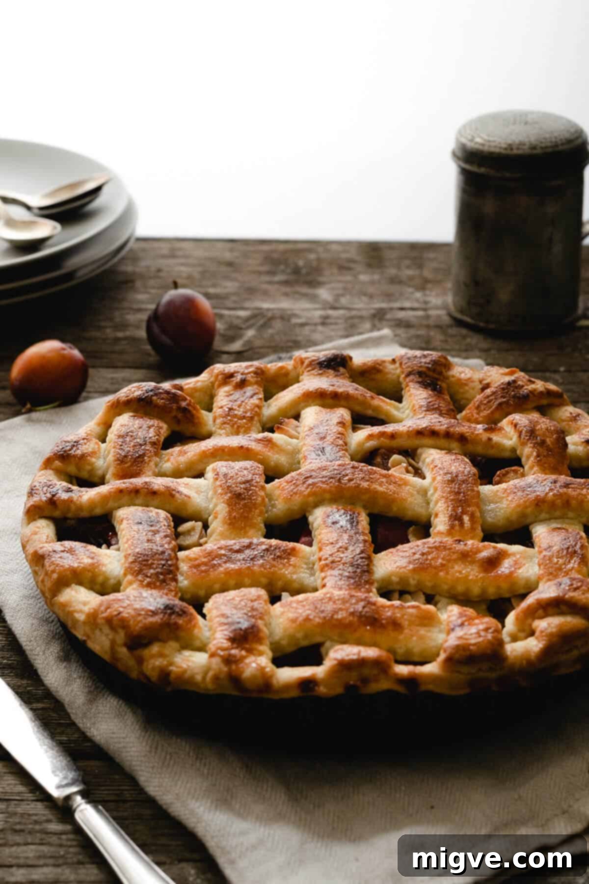 A baked plum and almond pie, golden brown and glistening, resting on a rustic kitchen table, inviting a slice.