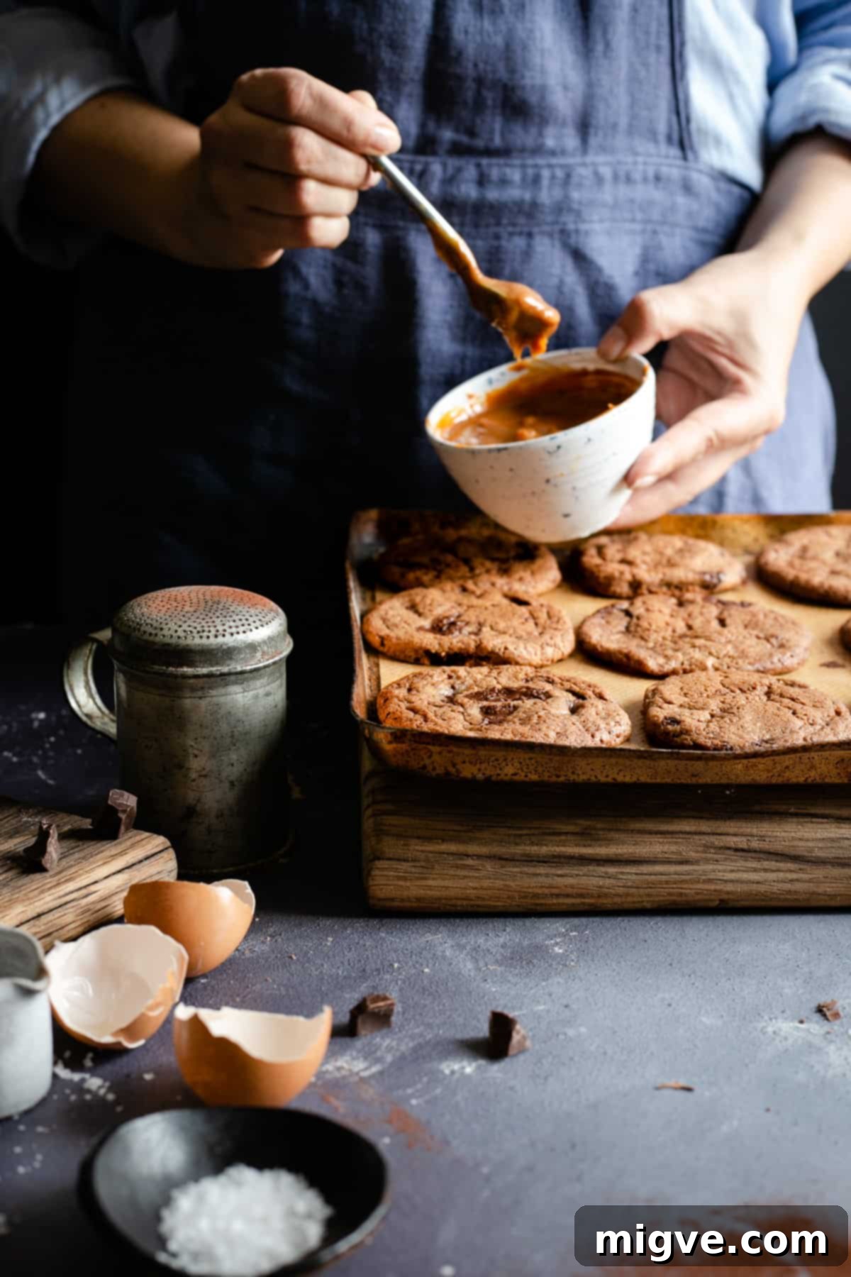 Decadent Chocolate and Salted Caramel Treats 2 A person holding a small bowl of luscious caramel above a tray laden with freshly baked, dark chocolate cookies, ready for topping.