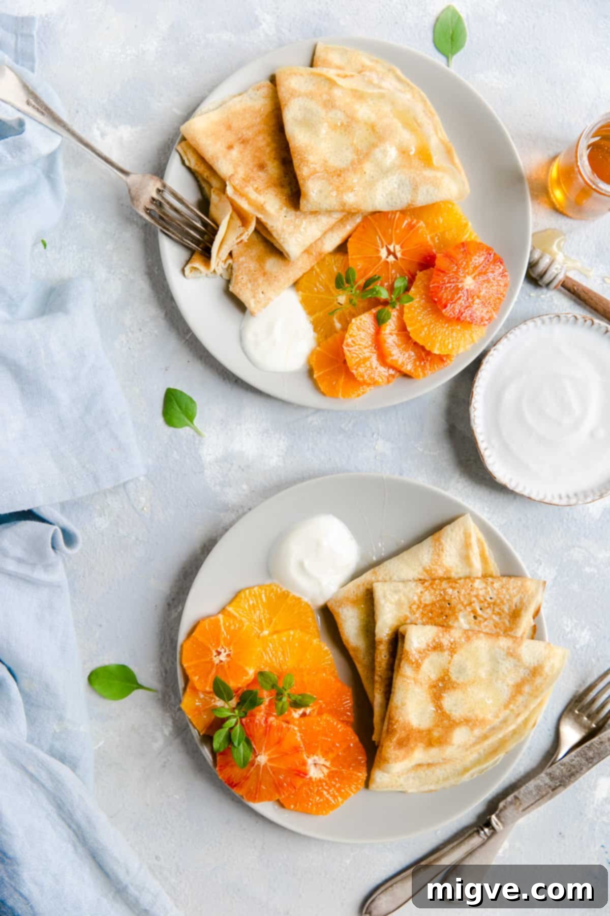 Overhead shot of two beautifully styled plates, each holding folded crepes, colorful citrus slices, creamy yogurt, and a tempting honey drizzle