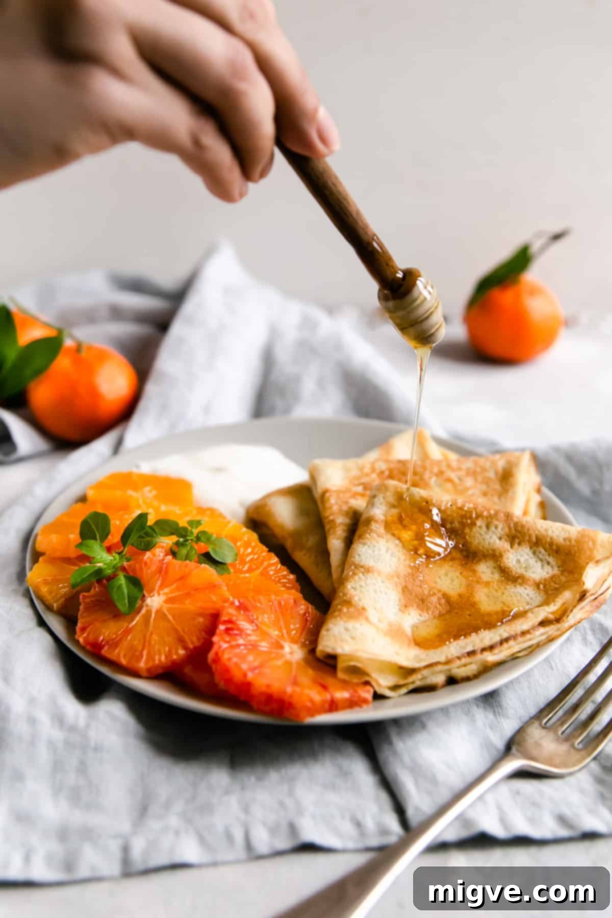 A person gently drizzling golden honey over a plate of freshly cooked crepes, vibrant citrus, and creamy yogurt