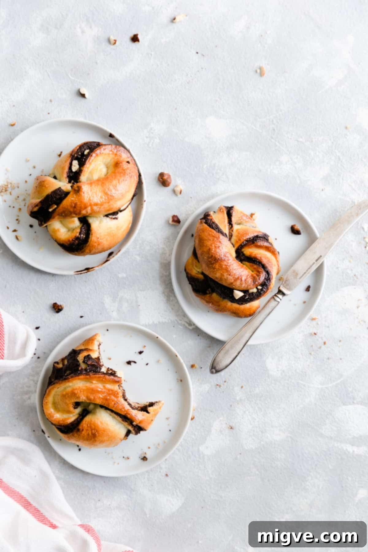 Swirled Chocolate Hazelnut Babka Buns 2 Overhead shot of 3 individual chocolate hazelnut babka buns resting on small white plates, showcasing their twisted layers and golden-brown crust.