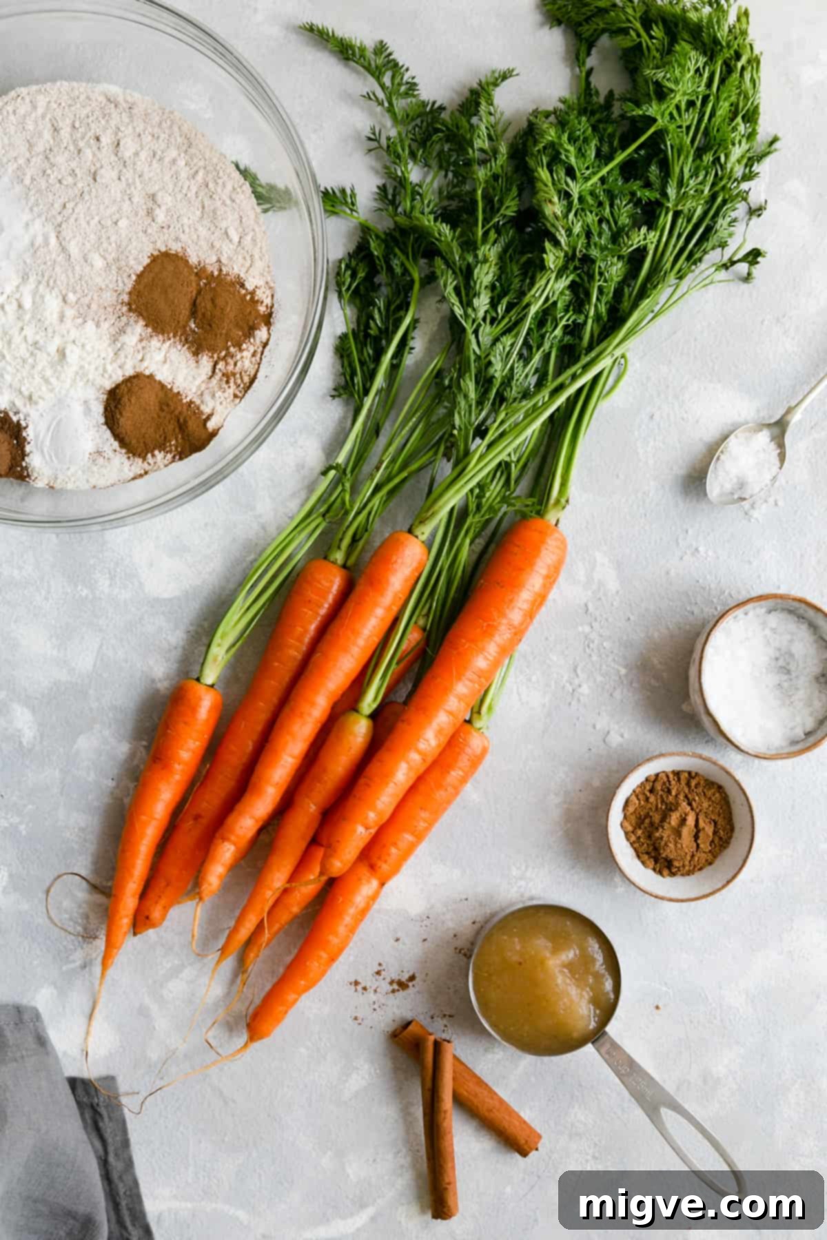 A vibrant flat lay shot displaying key vegan carrot cake ingredients: fresh carrots with their green tops, all-purpose flour, a jar of applesauce, and an assortment of aromatic spices.