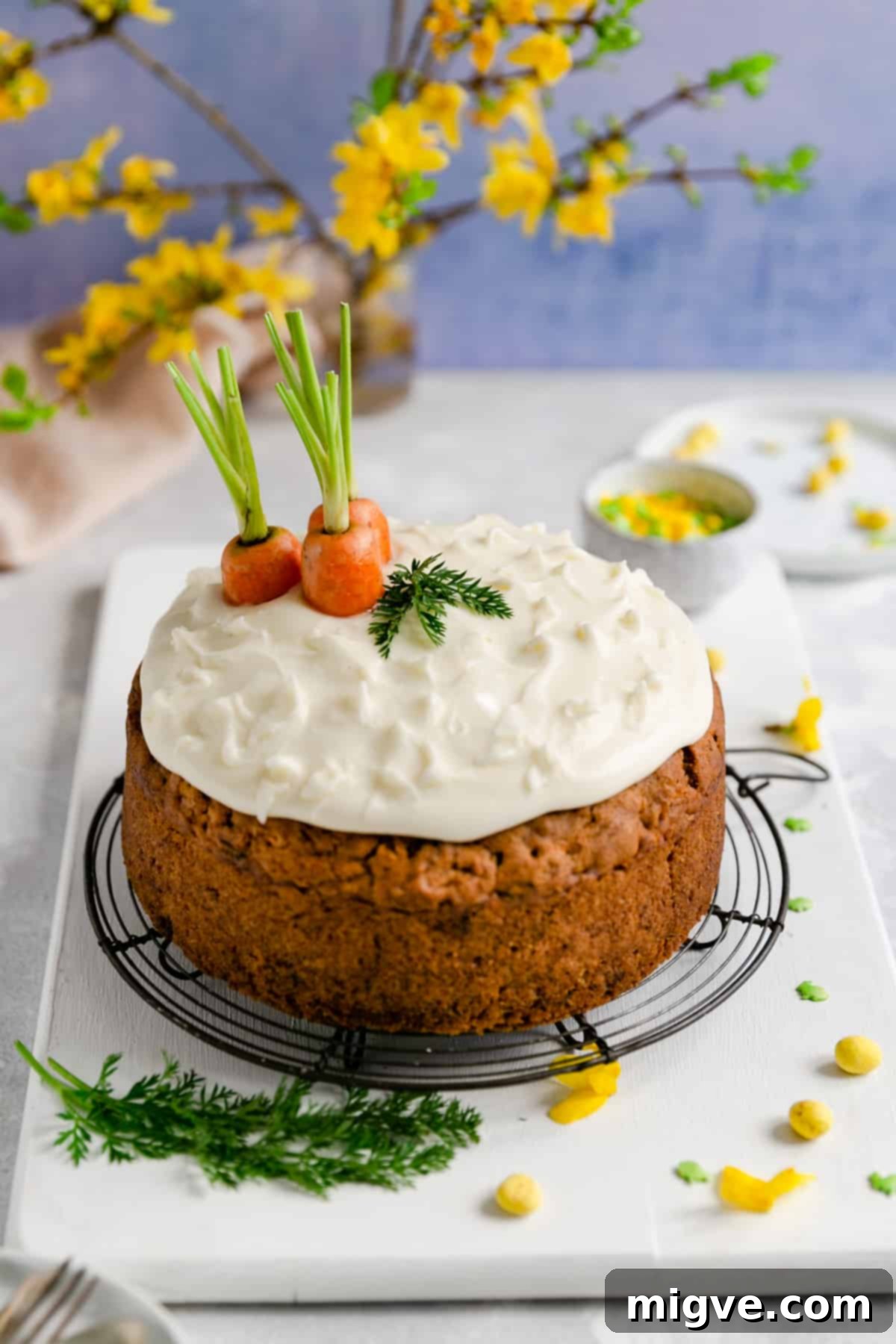 A delicious easy vegan carrot cake resting on a round metal cooling rack, with soft yellow flowers gently blurred in the background, creating a warm and inviting scene.
