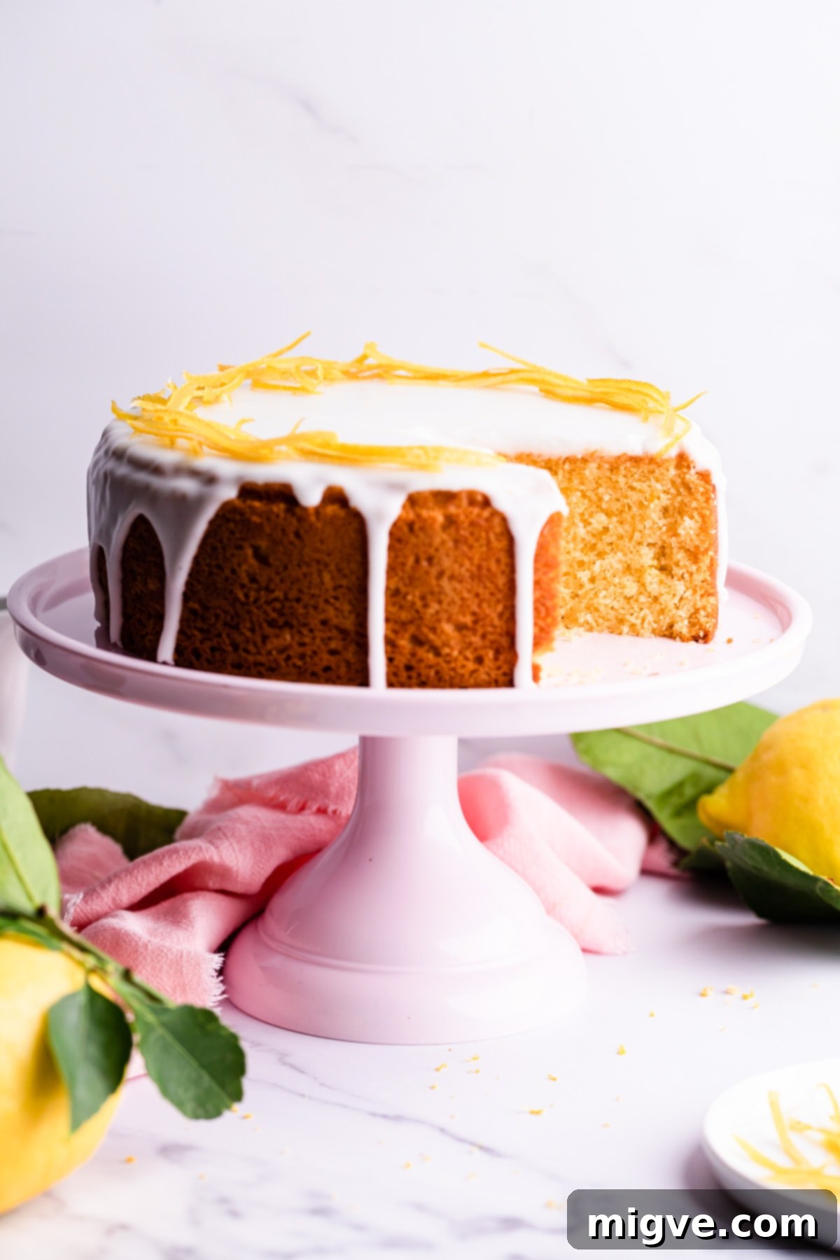 A vibrant lemon cake with a slice cut out, displayed on a pink cake stand with a pink napkin in the background.