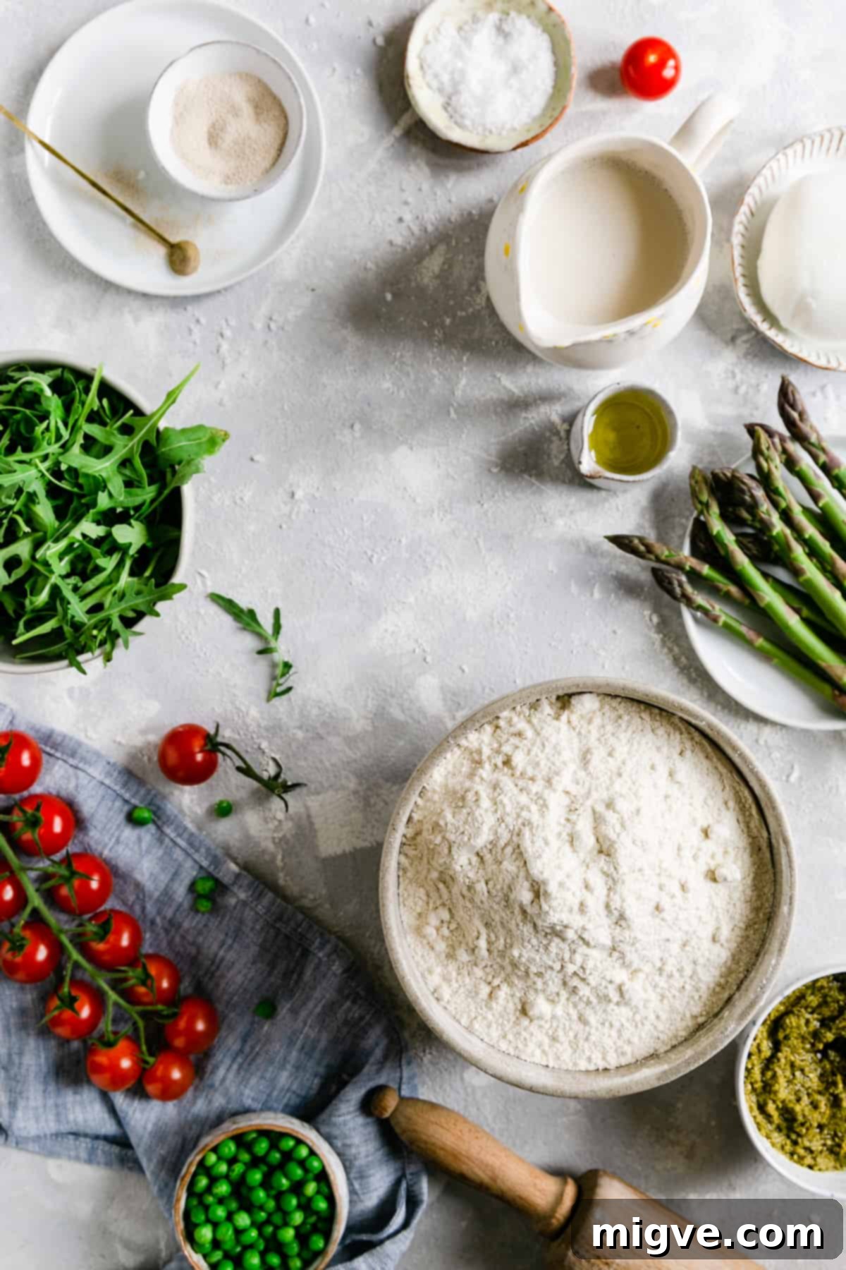 Overhead shot of all the fresh ingredients laid out for making the rocket and asparagus green pesto pizza, including flour, yeast, asparagus, peas, cherry tomatoes, mozzarella, and a jar of green pesto.