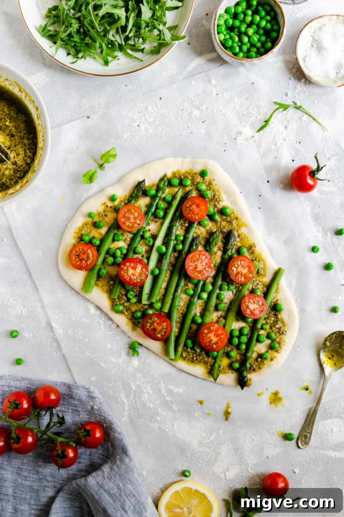 Top-down view of an unbaked pizza dough adorned with a generous layer of green pesto, carefully arranged blanched asparagus, bright green peas, and vibrant halved cherry tomatoes, ready for the oven.