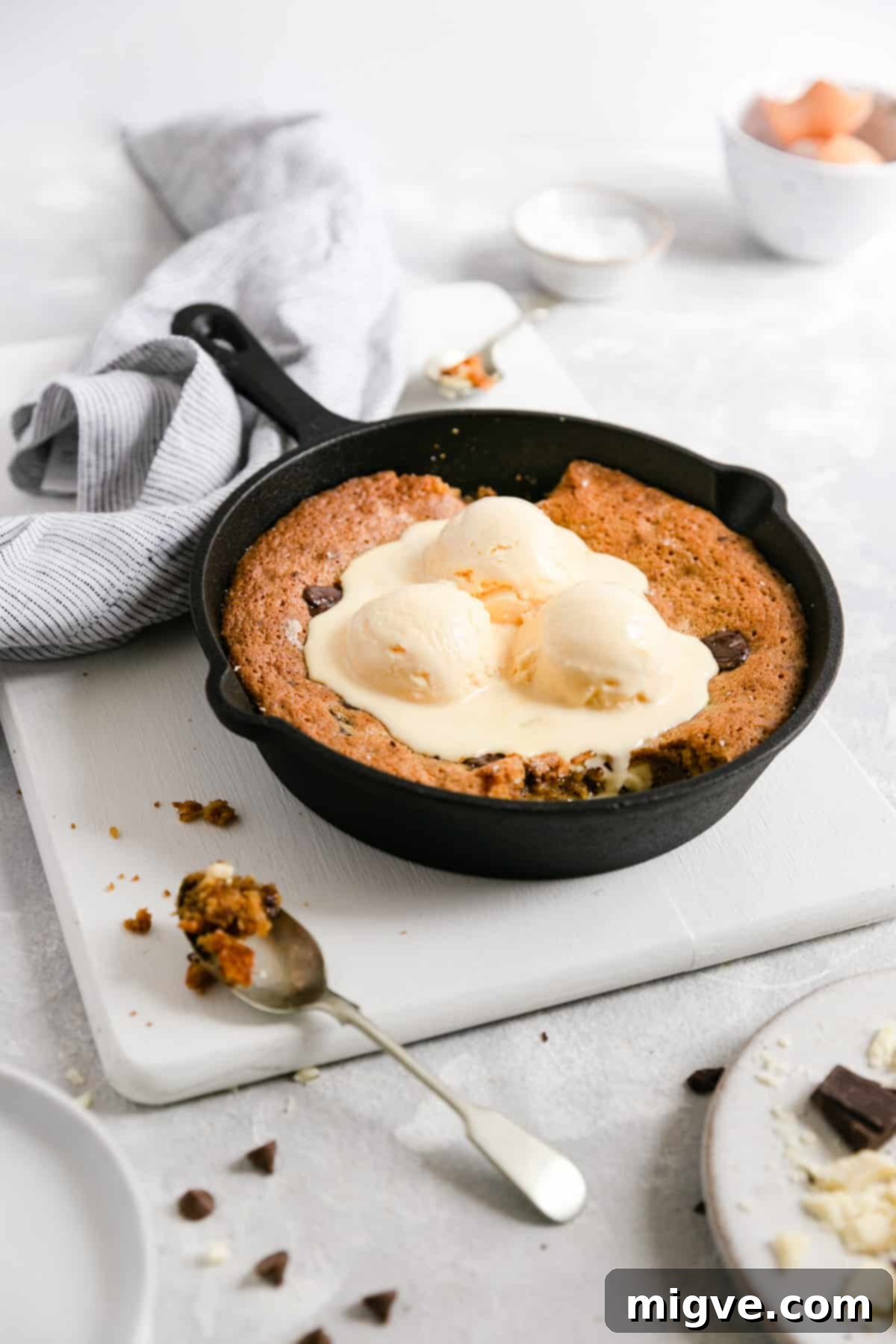 A side shot of a warm chocolate chip and malted milk cookie baking in a skillet, generously topped with creamy vanilla ice cream, ready to be enjoyed.