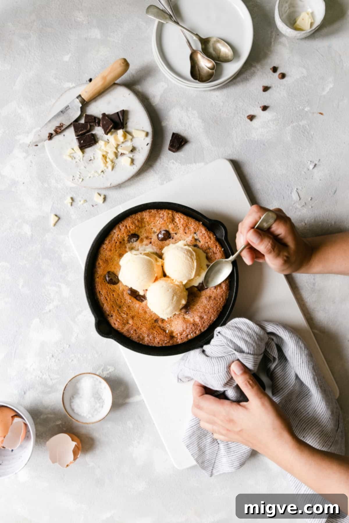 Top view of a hand holding a small skillet containing a warm chocolate chip cookie, gloriously topped with scoops of melting vanilla ice cream, promising an irresistible dessert.