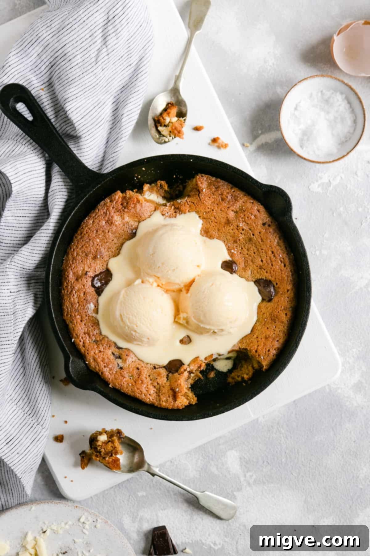 Overhead shot of a rich chocolate chip and malted milk cookie in a skillet, with a spoon already having scooped out a generous portion, revealing its gooey interior.