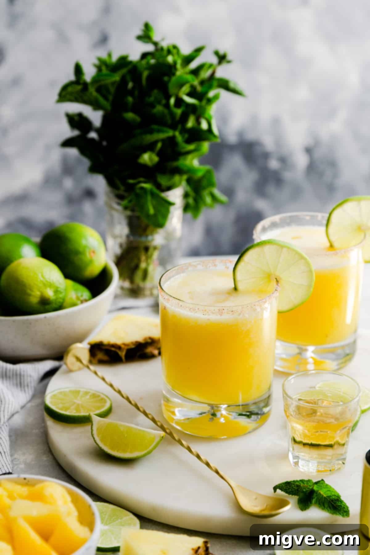 Two frosty glasses of frozen pineapple margaritas garnished with lime and mint on a marble board, ready to serve.