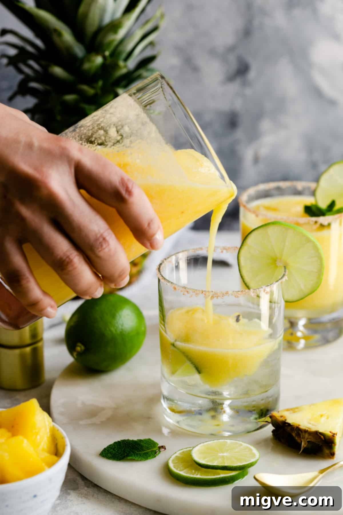 A hand gently pouring a vibrant yellow frozen pineapple margarita from a blender into a stemmed glass, emphasizing the smooth, icy texture.