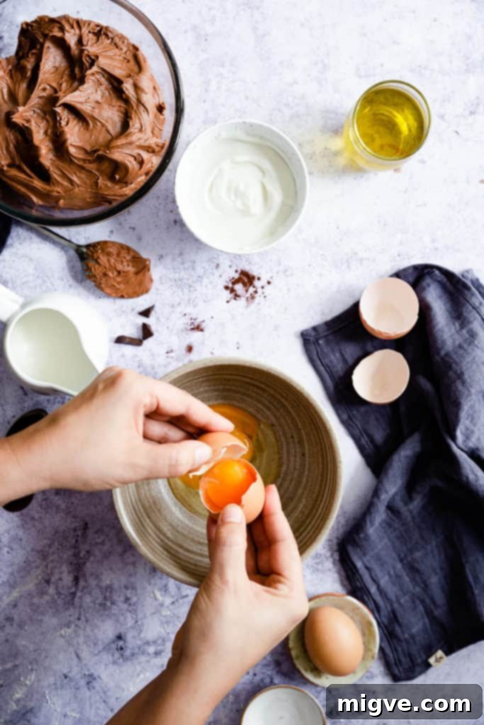 Top view of hands cracking eggs into a mixing bowl, part of the wet ingredient preparation for the chocolate cake.