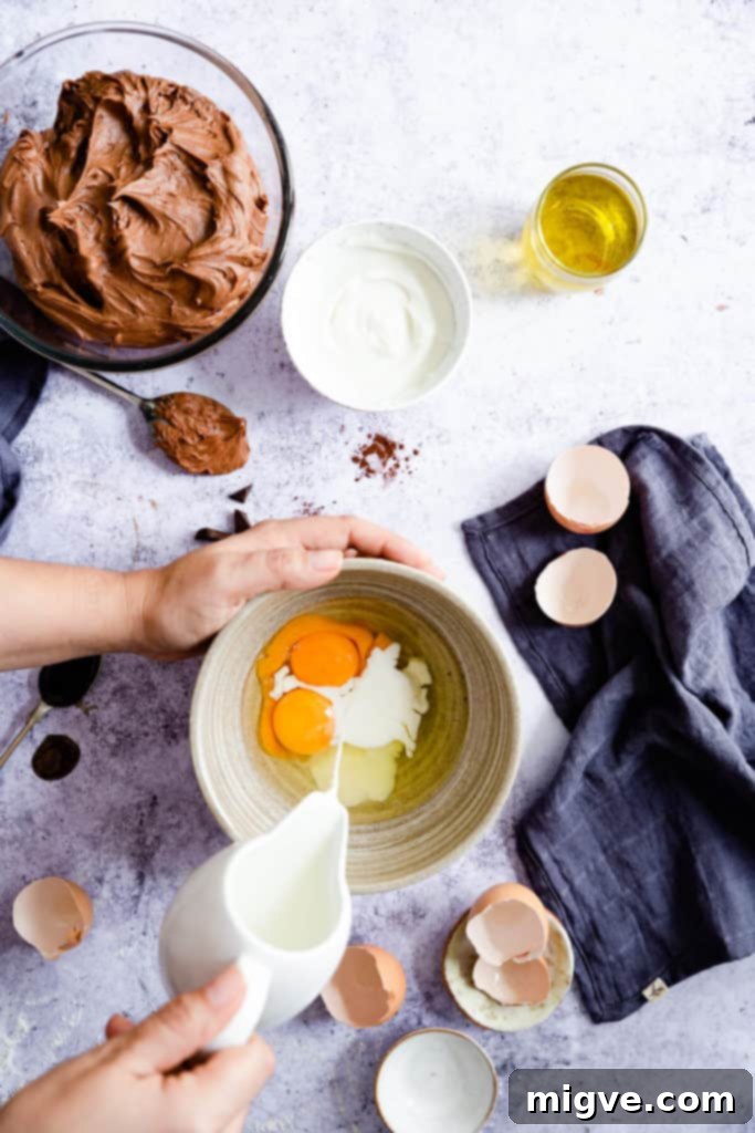 Top view of hands pouring milk into a bowl containing whisked eggs, contributing to the wet ingredients for the cake batter.