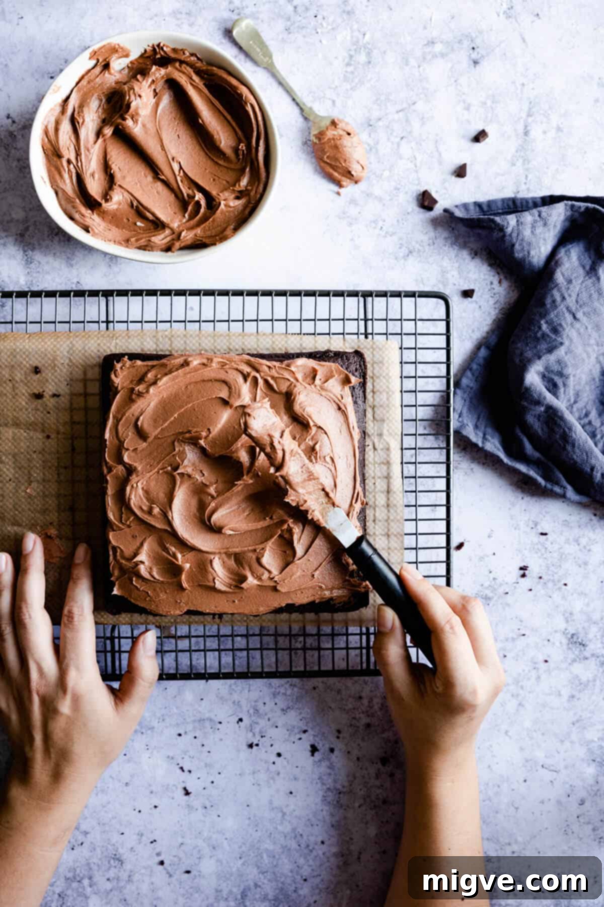 Overhead shot of hands gracefully spreading smooth chocolate buttercream over a freshly baked dark chocolate traybake.