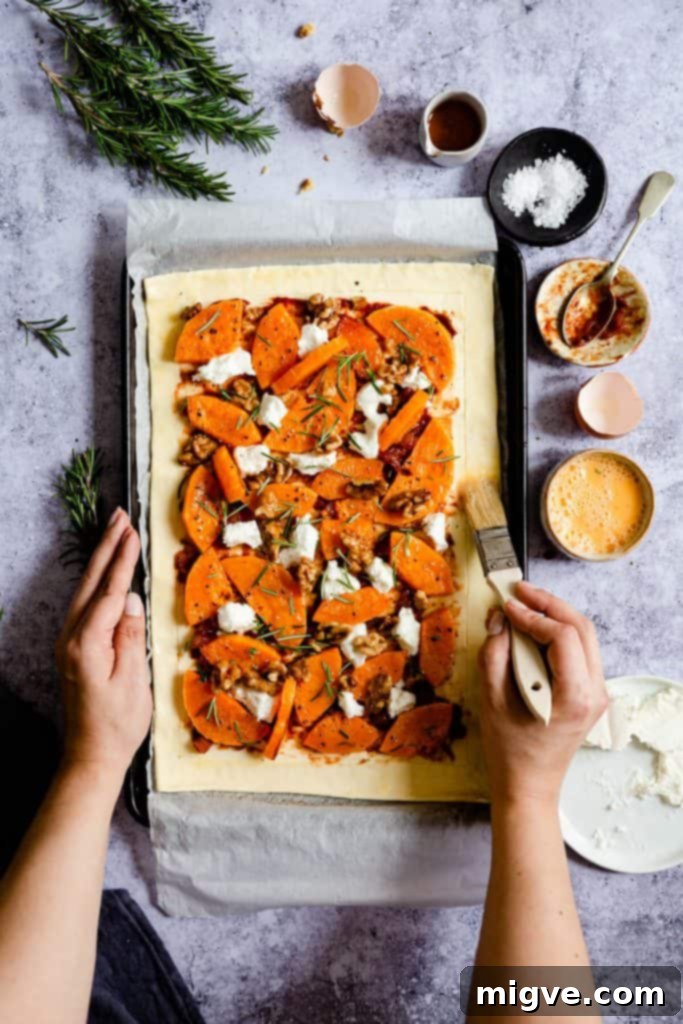 Top-down view of a hand brushing the scored puff pastry border with a golden egg wash, preparing it for a beautiful, crispy finish.