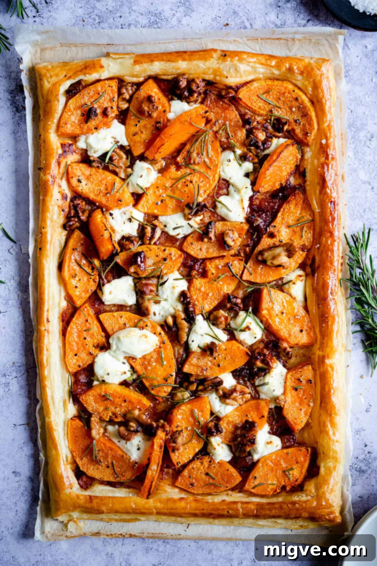 Top-down shot of a whole savoury tart, showcasing the vibrant colours and textures of goats' cheese, roasted butternut squash, and fresh rosemary sprigs atop a golden puff pastry base.