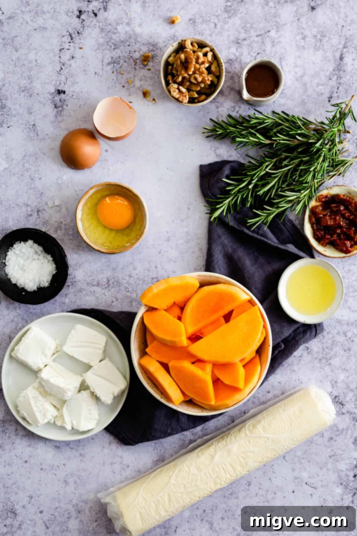 Overhead view of various fresh ingredients neatly arranged in bowls on a rustic wooden surface, including chopped butternut squash, walnuts, goats' cheese, and fresh rosemary, ready for the tart recipe.