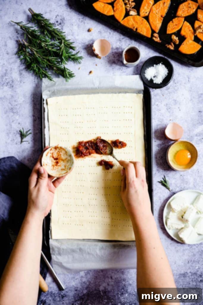 Overhead shot of hands spreading a rich red tomato chutney evenly across the surface of the puff pastry, staying within the scored border.