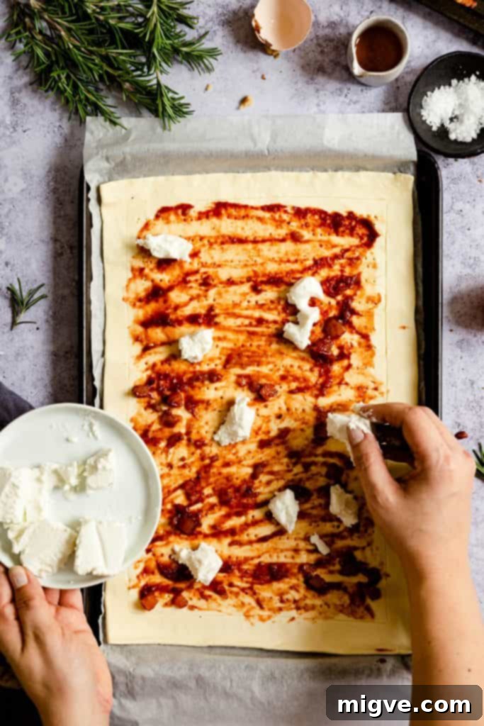 Top-down view of small, creamy chunks of goats' cheese being casually scattered over the chutney-covered puff pastry base of the tart.