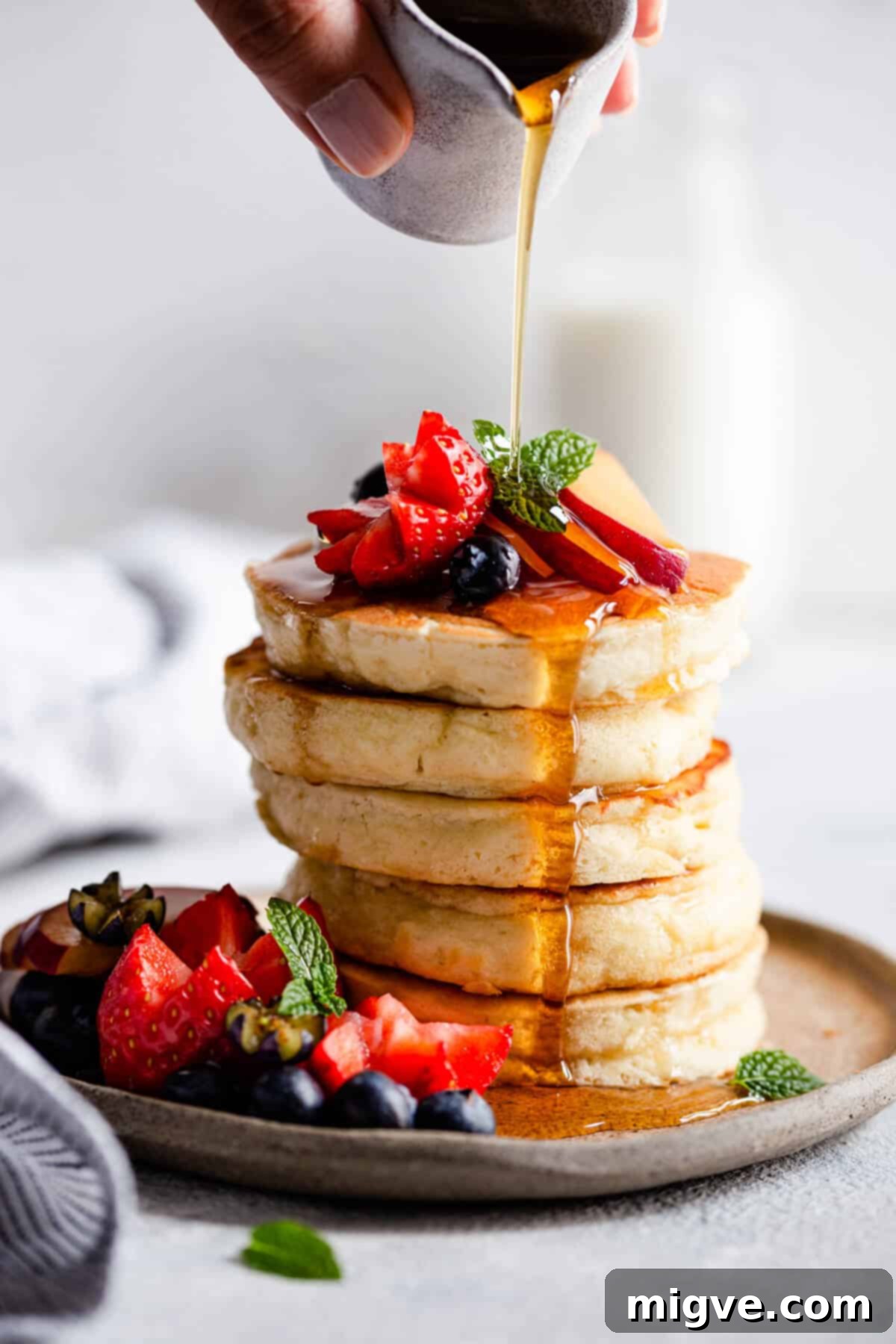 A close-up side view of golden fluffy pancakes being generously drenched with maple syrup.