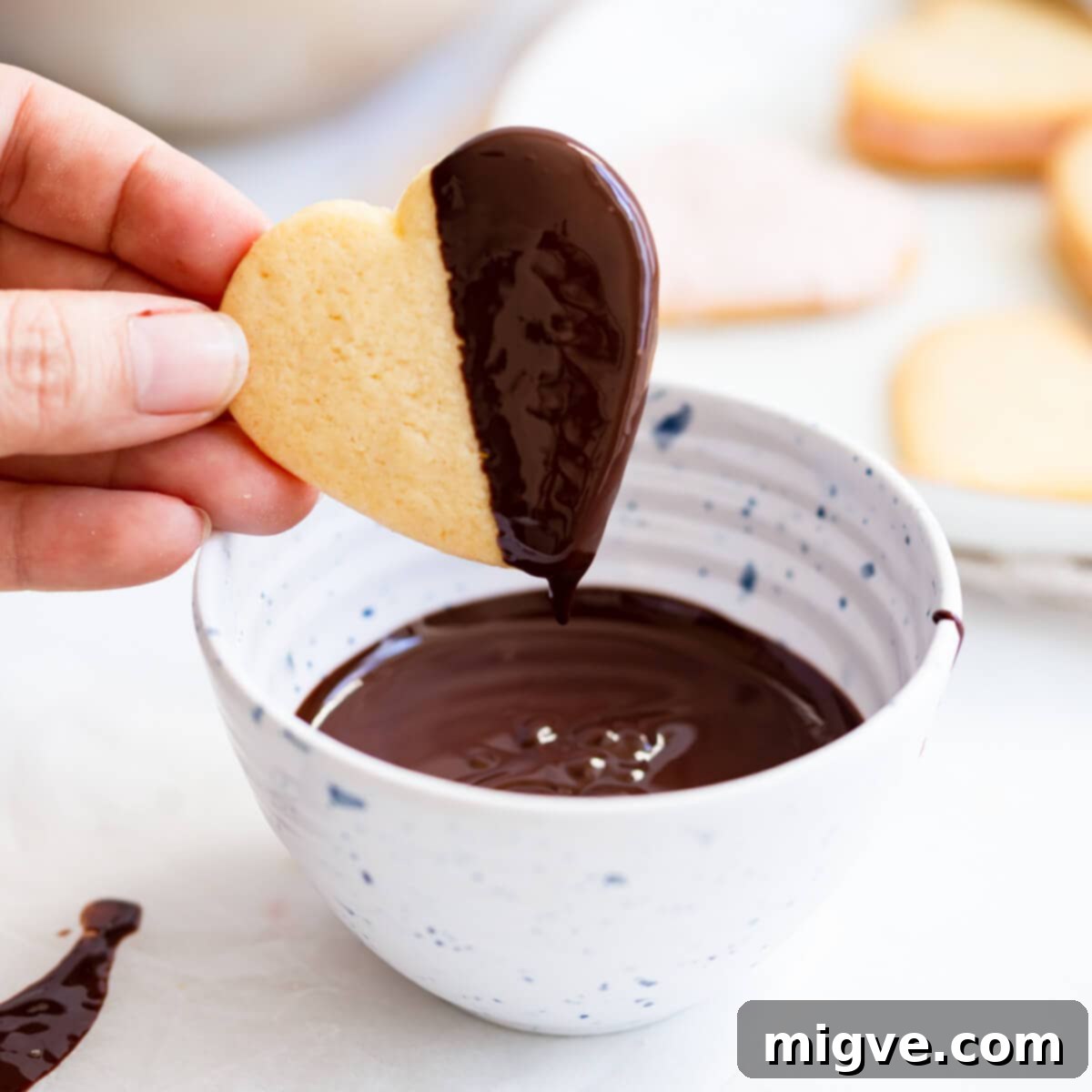 Sweetheart Sugar Cookies 7 A heart-shaped sugar cookie being dipped into a bowl of glossy melted chocolate.