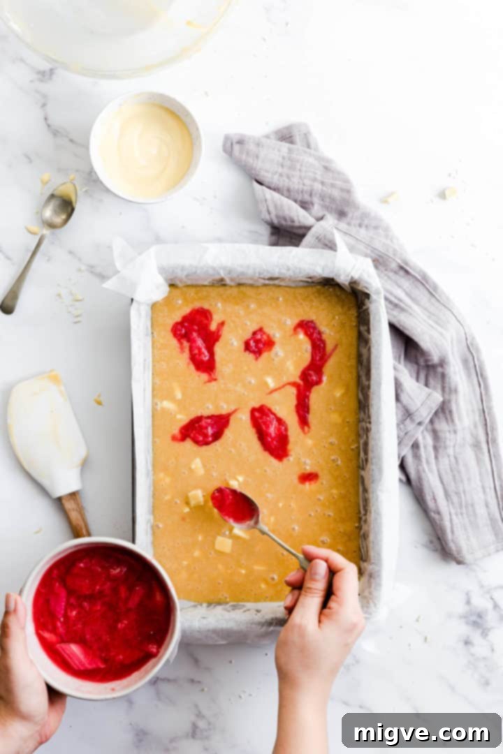 Top view of a person carefully adding dollops of vibrant rhubarb compote onto the smooth blondie batter spread evenly in a baking tin.