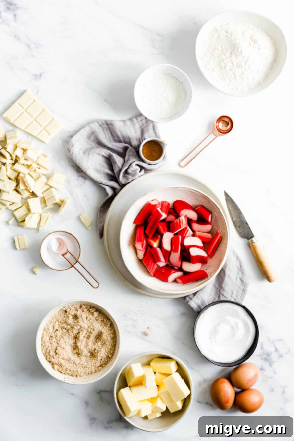 Overhead shot showcasing all the fresh and dry ingredients laid out for making rhubarb and custard blondies, including butter, sugars, eggs, rhubarb, and white chocolate.