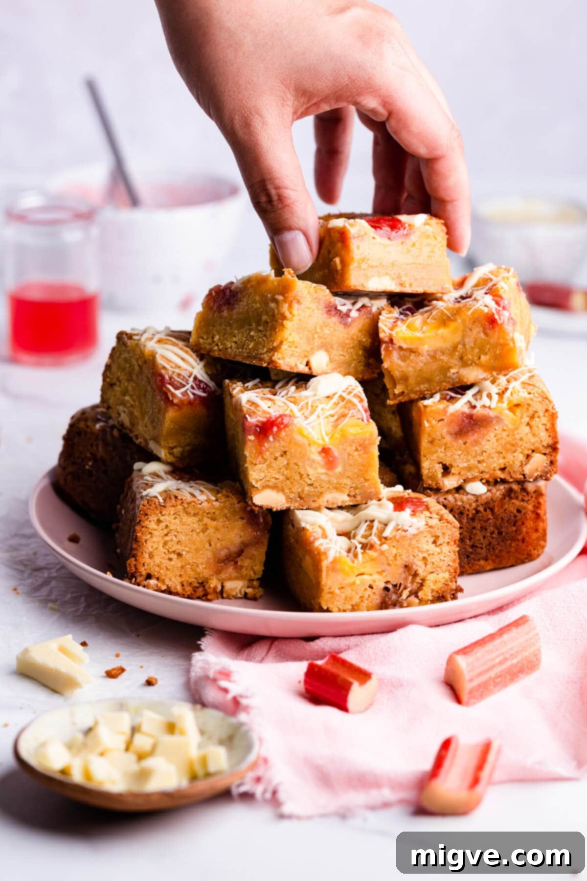 Side close-up of a plate filled with freshly cut rhubarb blondie squares, with a hand reaching to take a piece, highlighting their irresistible nature.