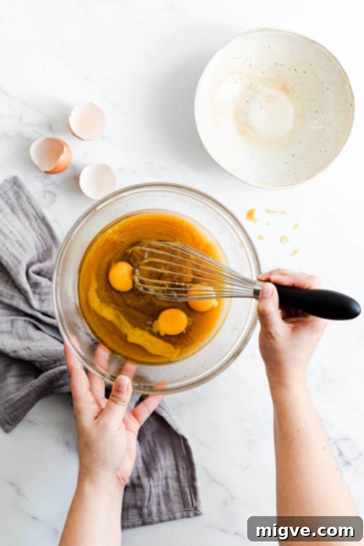 Overhead shot of a person gently mixing room temperature eggs into the cooled butter and sugar batter in a large mixing bowl.