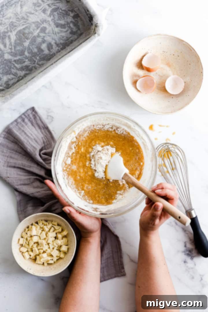 Top view of a person stirring dry ingredients like flour, custard powder, and baking powder into the prepared blondie cake batter in a mixing bowl.
