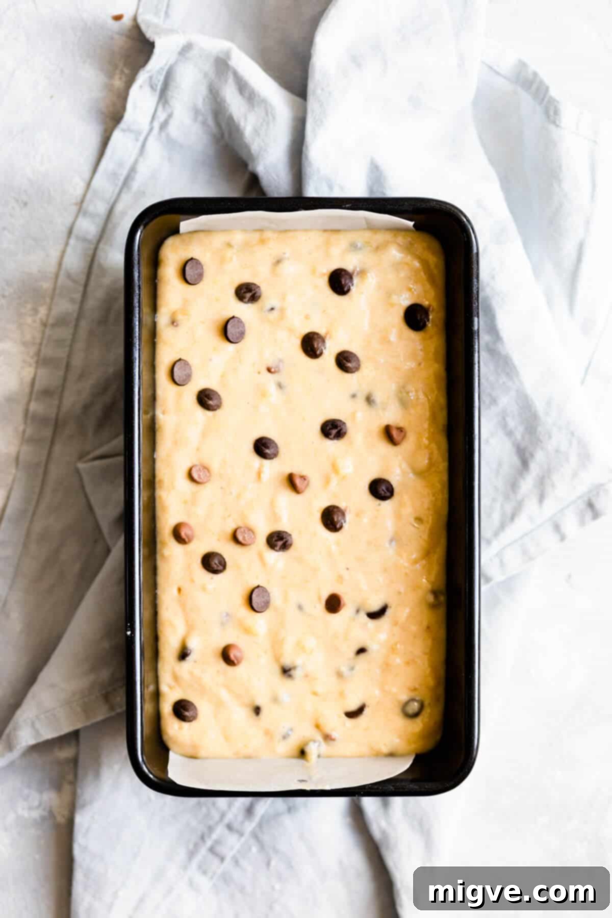 Overhead shot of a loaf tin filled with smooth chocolate chip banana bread batter, with a sprinkle of extra chocolate chips on top, ready to be baked.