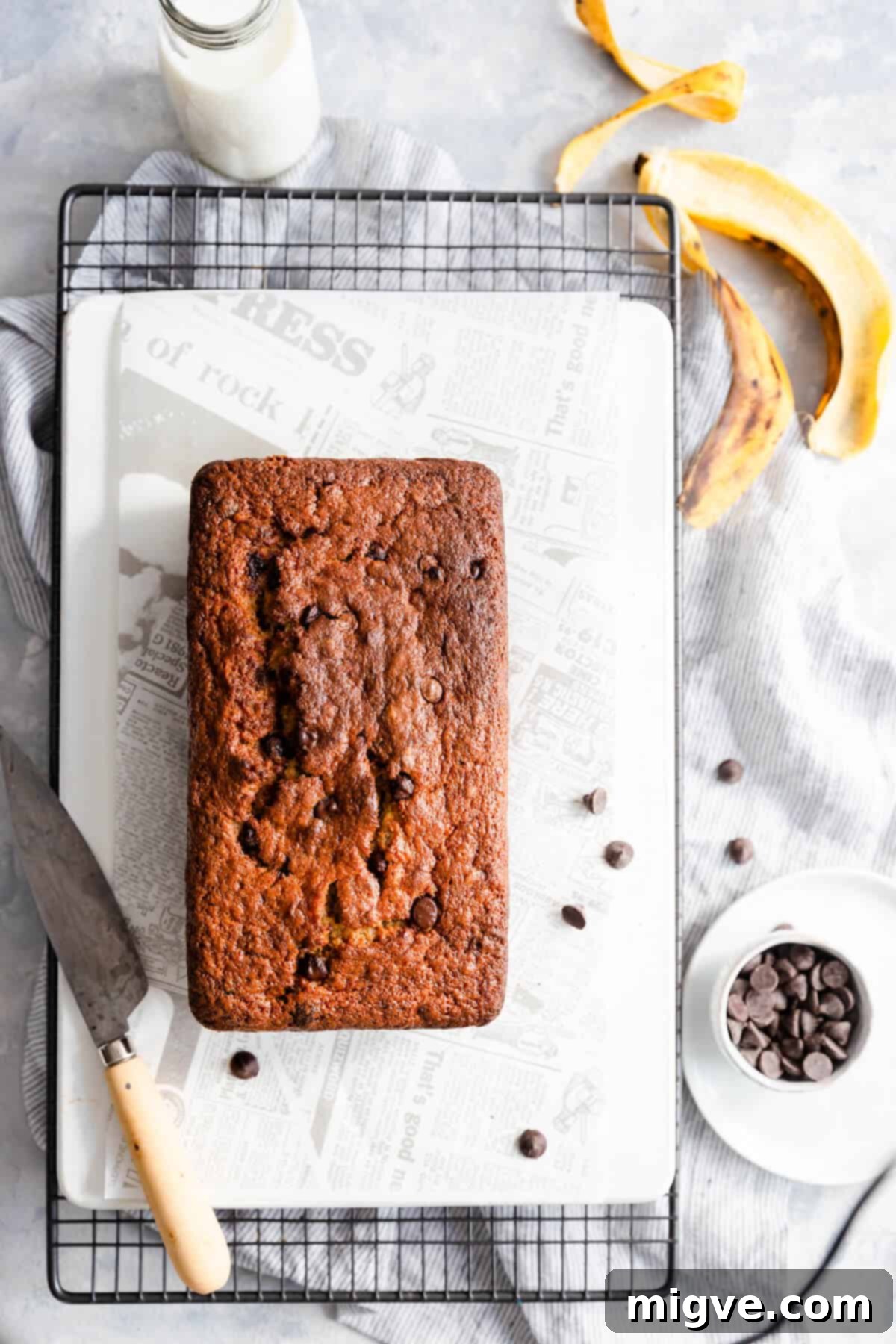 Overhead view of a beautifully golden-brown chocolate chip banana bread loaf on a white cutting board, revealing the scattered chocolate chips on top.