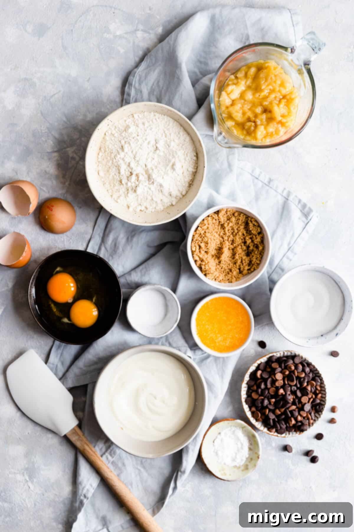 Overhead view of various baking ingredients, including flour, sugar, butter, and ripe bananas, neatly arranged in bowls on a rustic wooden surface, ready for making banana bread.