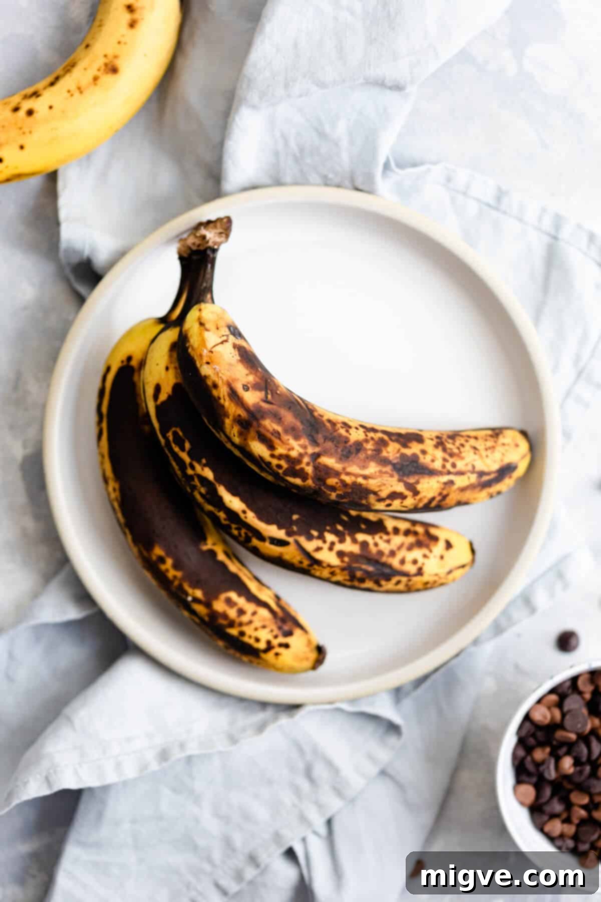 Top-down view of three perfectly ripe, spotty bananas on a white plate, ready for baking.