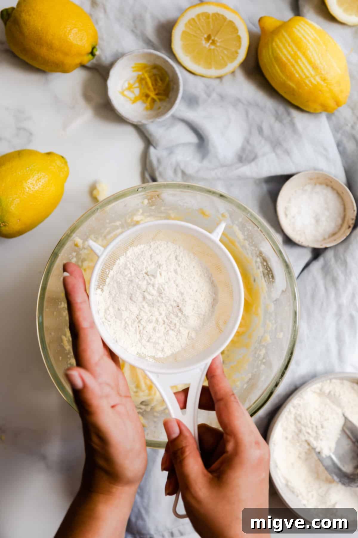 Zesty Small Batch Lemon Drizzle Cake 10 Overhead shot of a person carefully sieving self-rising flour and salt into the cake batter in a bowl.