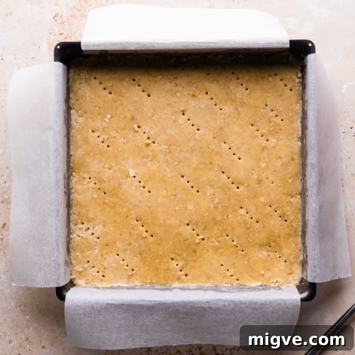 A square baking tin, meticulously lined with baking parchment, with ⅔ of the crumble dough pressed into its base and gently pricked with a fork.