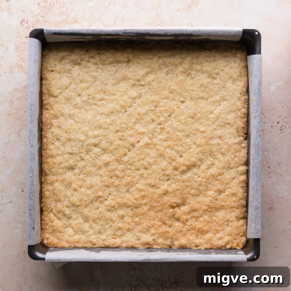 A square baking tin, lined with parchment paper, containing the perfectly baked golden-brown crumble base.