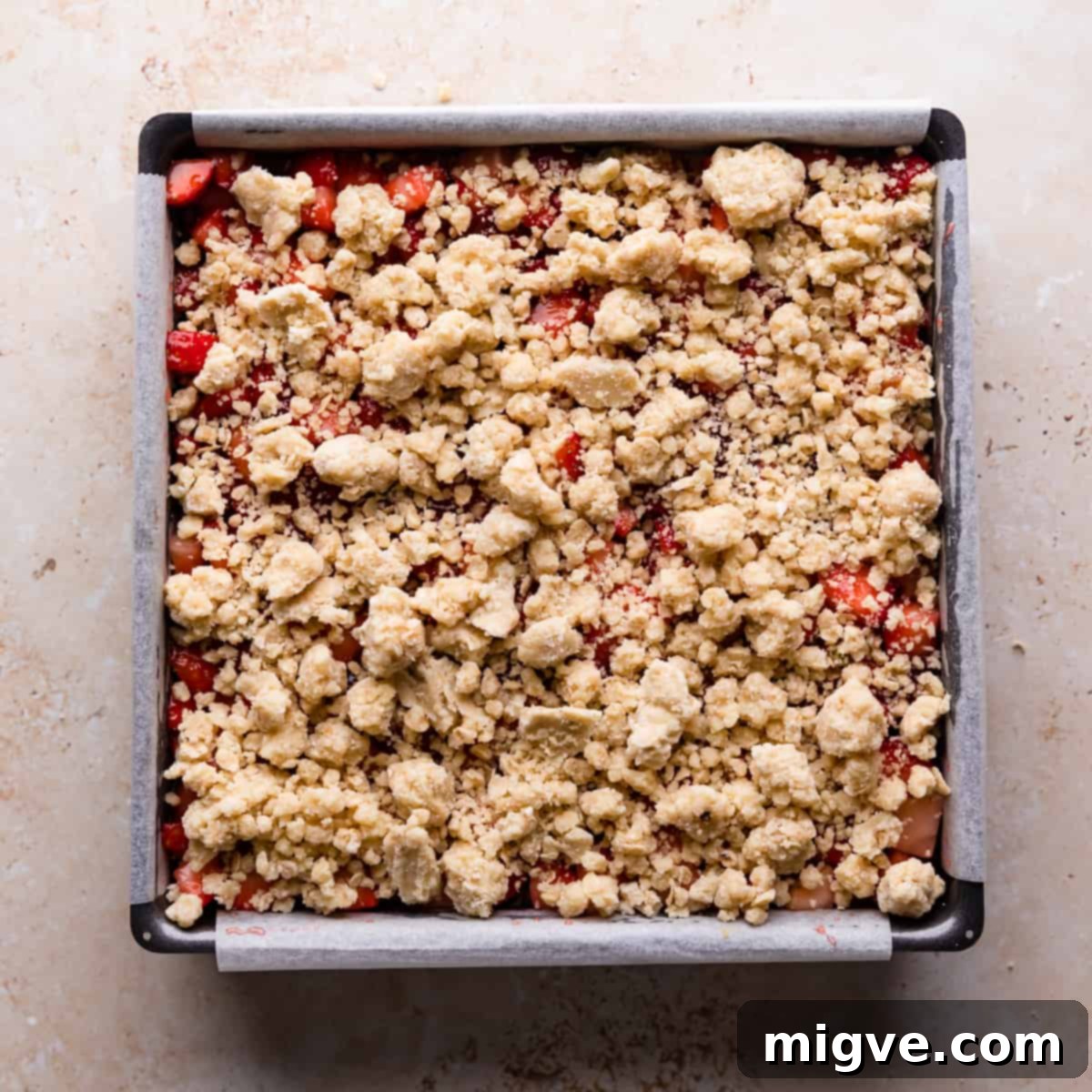 A square baking tin filled with the strawberry layer, now generously topped with the remaining crumble mixture, ready for its final bake.