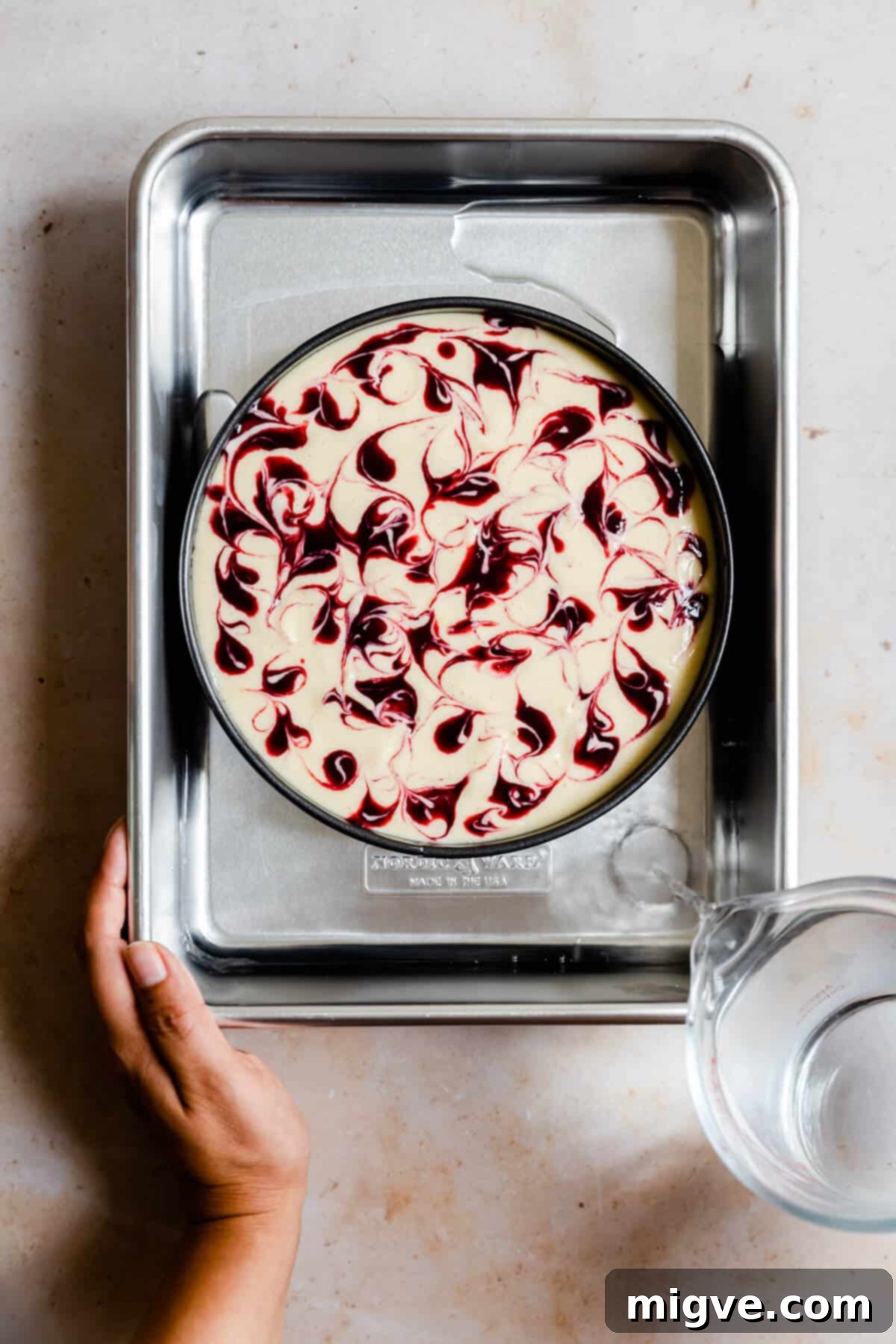 Overhead shot of a person carefully pouring water into a large roasting pan, creating a water bath around a cheesecake tin.