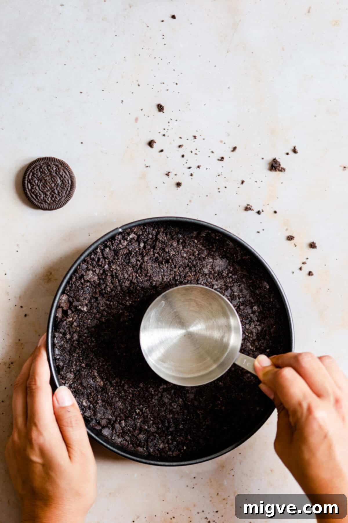 Top view of a person firmly pressing an Oreo cookie crust into the bottom of a baking tin using a measuring cup.
