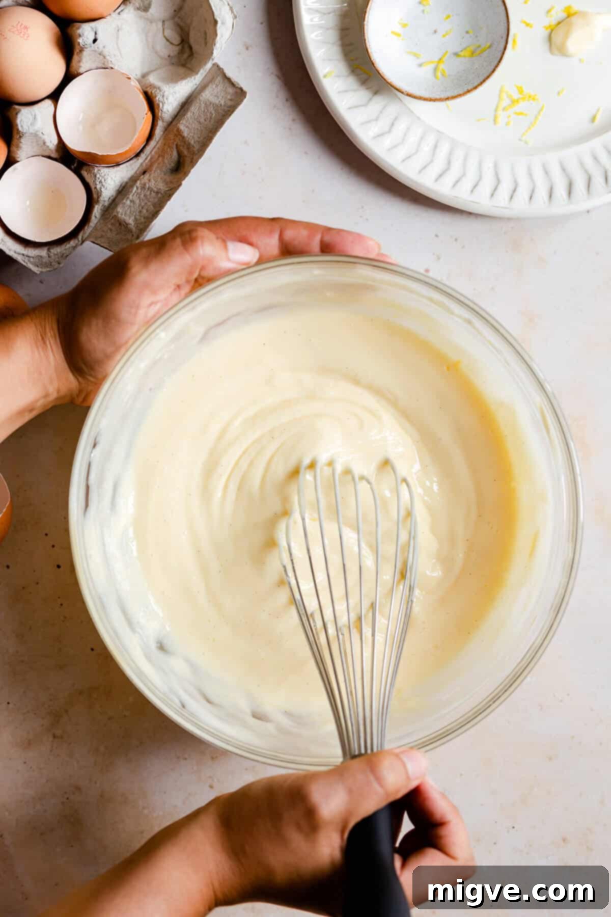Top view of a person gently whisking smooth cheesecake batter in a large mixing bowl.