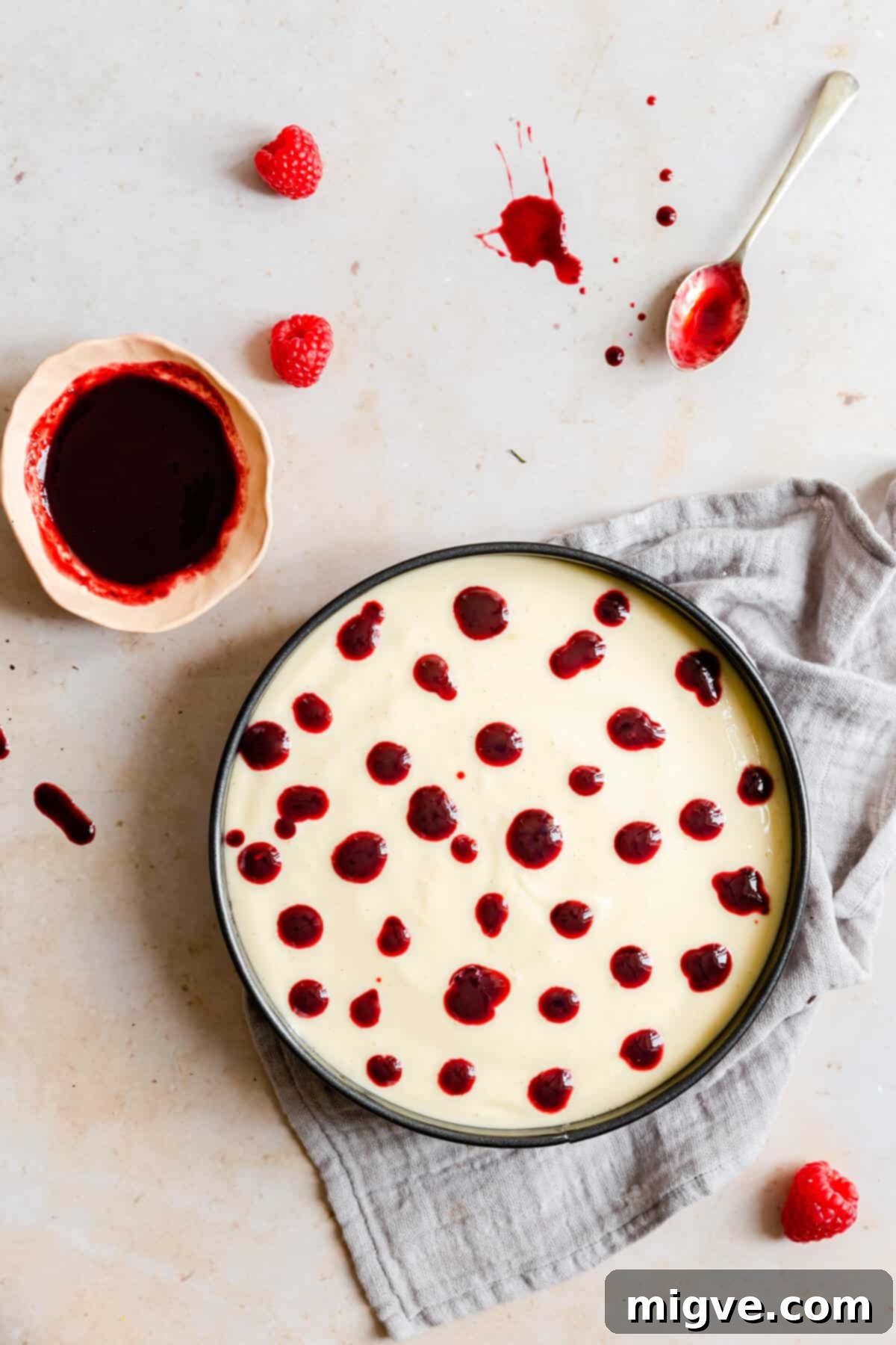 Overhead shot of cheesecake batter poured into a baking tin, topped with dollops of vibrant raspberry sauce.