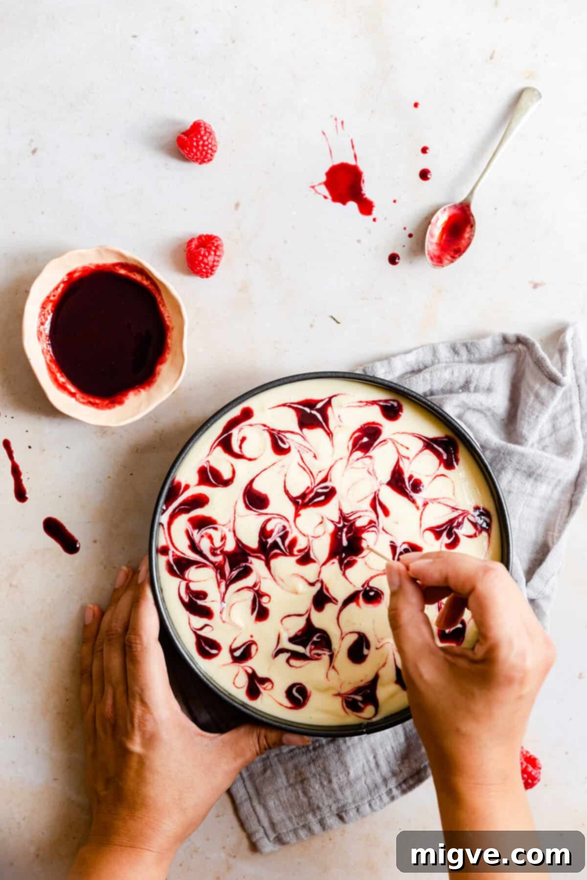 Top view of a person creating beautiful swirl patterns in the cheesecake batter with a toothpick after adding raspberry sauce.