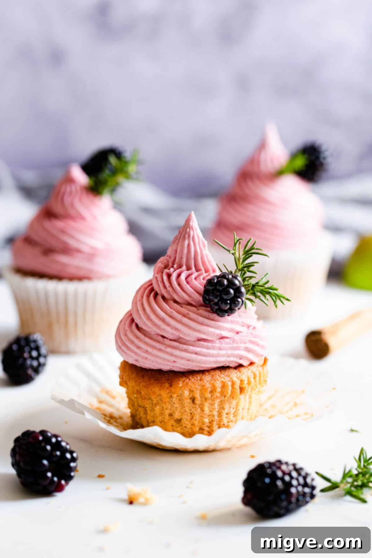 Straight ahead shot of a spiced apple and blackberry cupcake topped with fresh blackberry and a sprig of rosemary, showcasing the fluffy cake and vibrant pink buttercream.