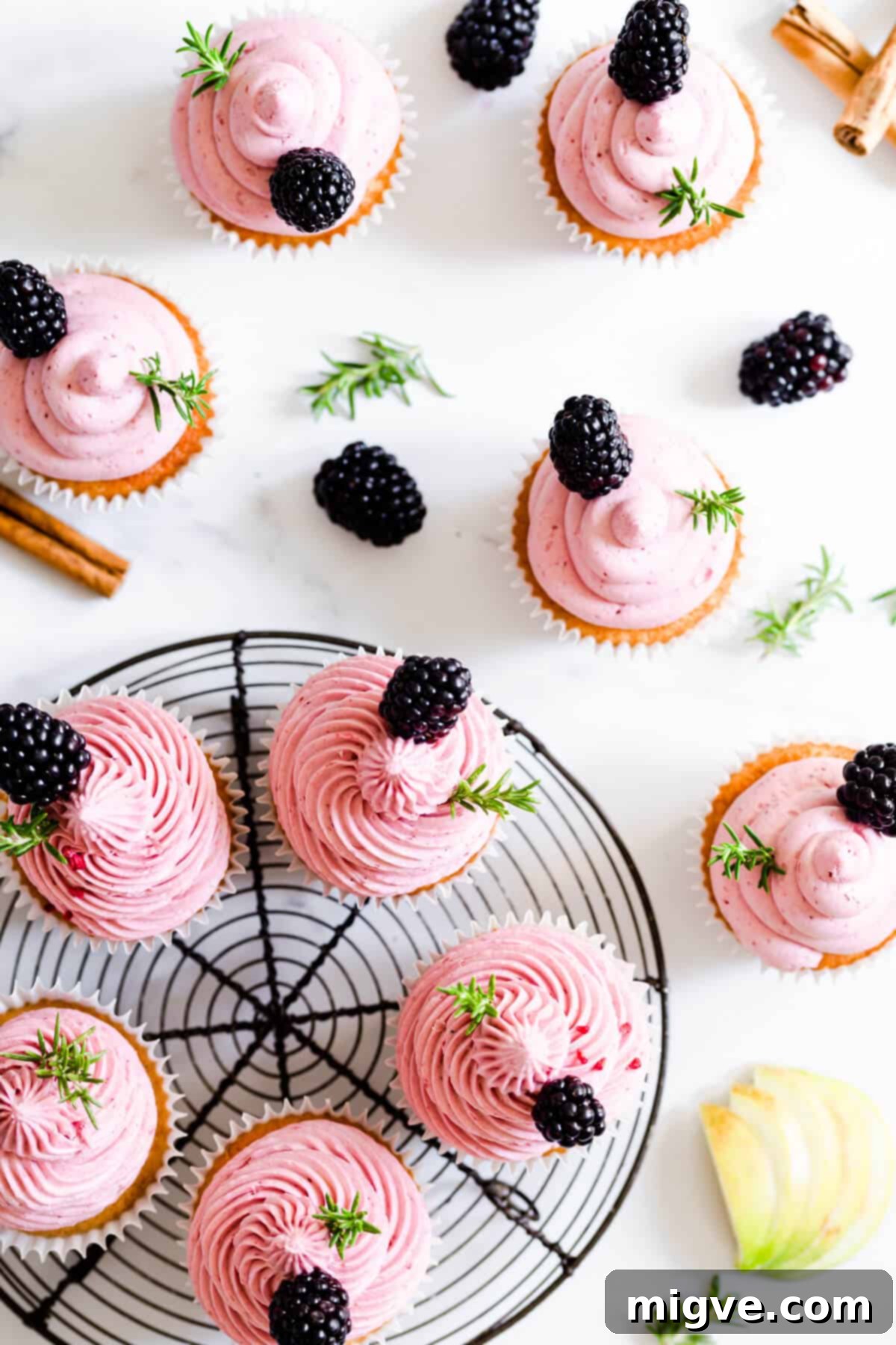 Overhead shot of freshly baked spiced apple cupcakes, each generously topped with pink blackberry buttercream and garnished with fresh rosemary.