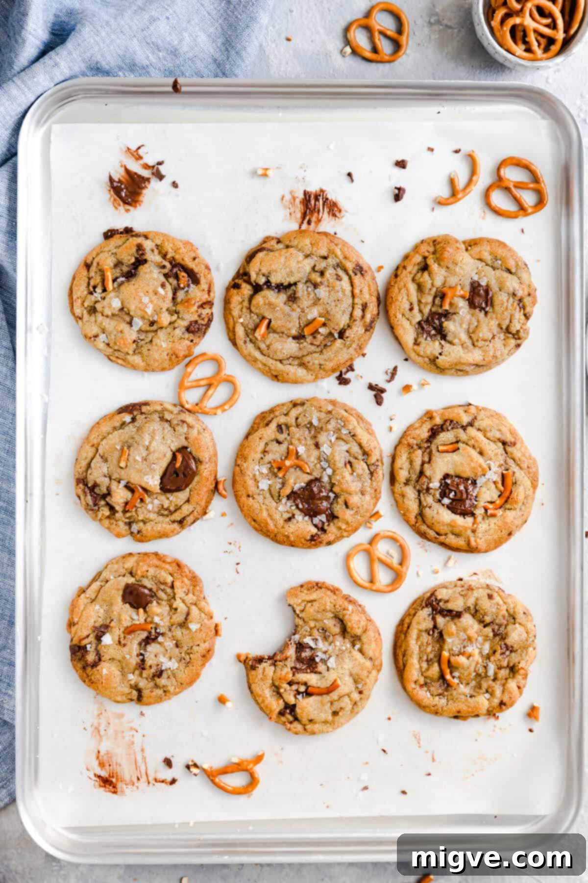 Overhead shot of a baking tray filled with nine perfectly golden chocolate pretzel cookies, topped with visible pretzel pieces and melted chocolate, with one cookie showing a bite taken out.