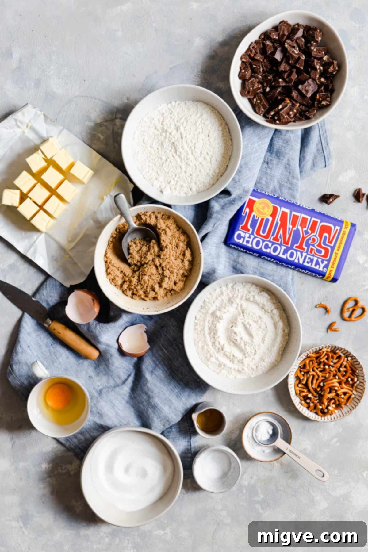 Overhead shot of various ingredients for chocolate chip pretzel cookies neatly arranged in bowls on a white surface.