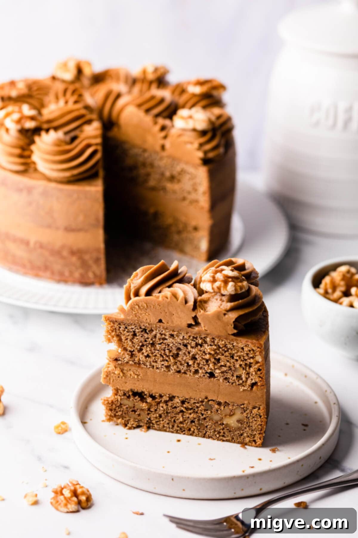 slice of coffee walnut cake on a plate with the rest of the cake in the background.