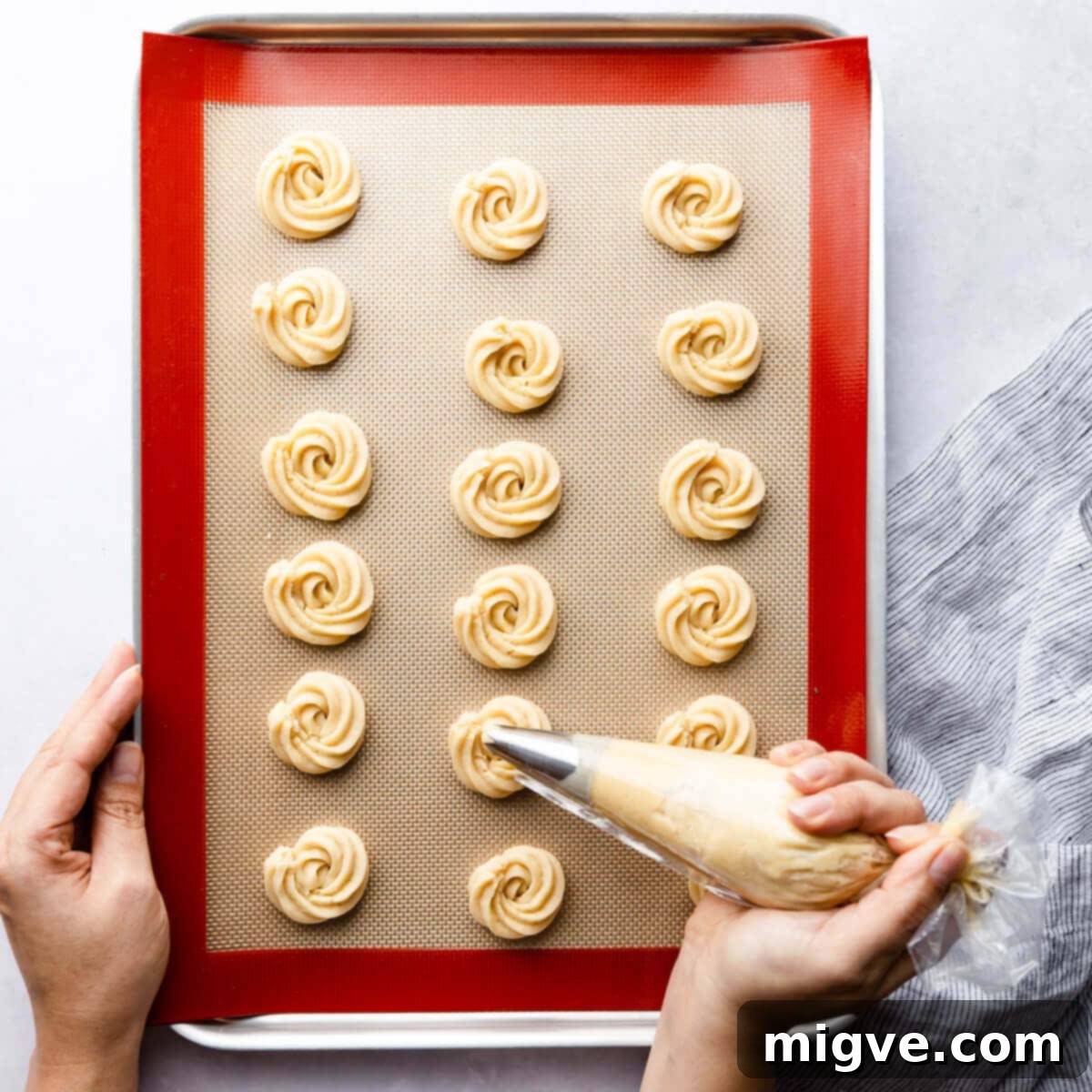 Buttery Danish Cookie Perfection 6 A person expertly piping cookie dough into intricate shapes onto a baking sheet lined with a silicone mat.